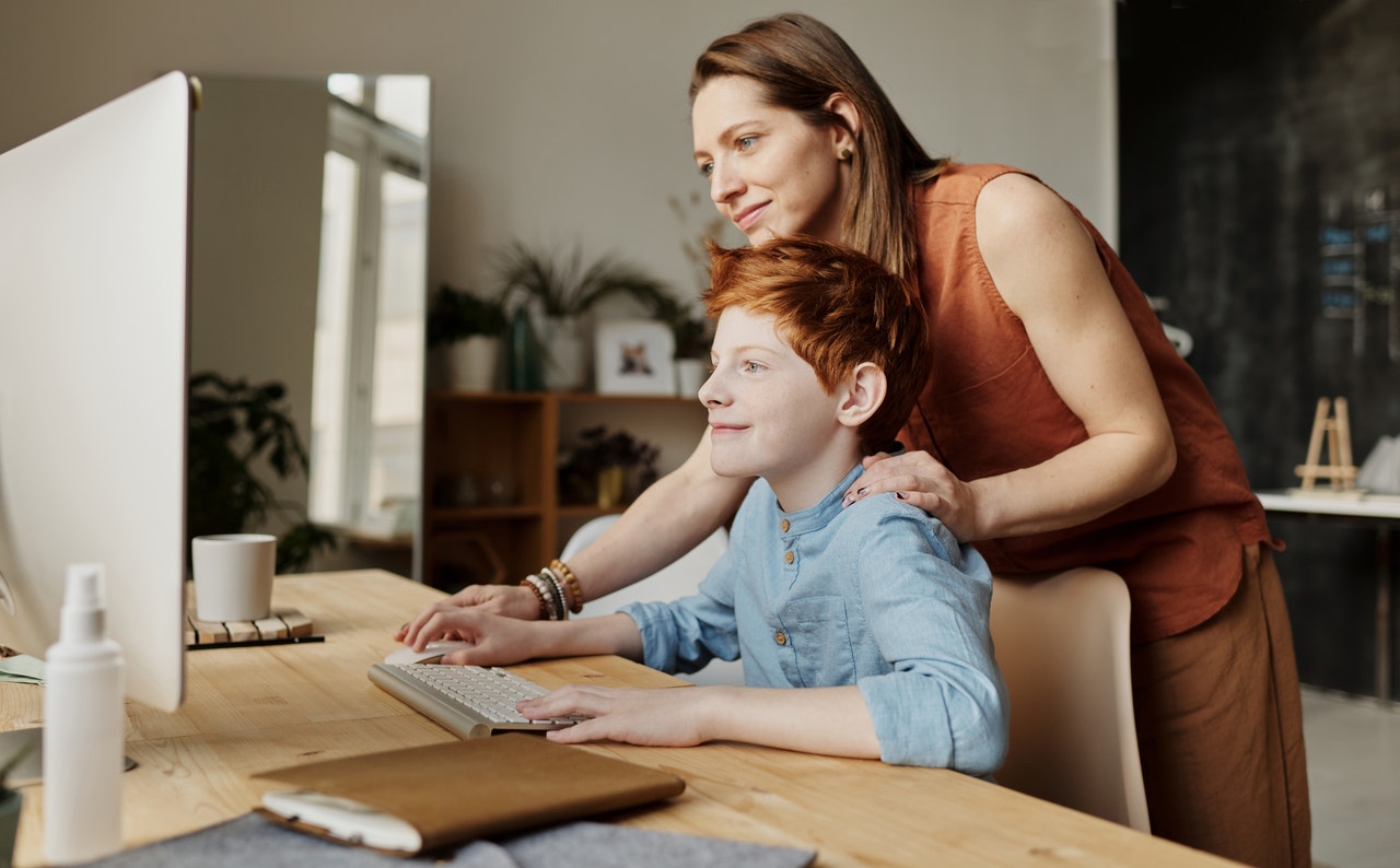 A woman guides her young son while he uses the computer, They are both smiling.