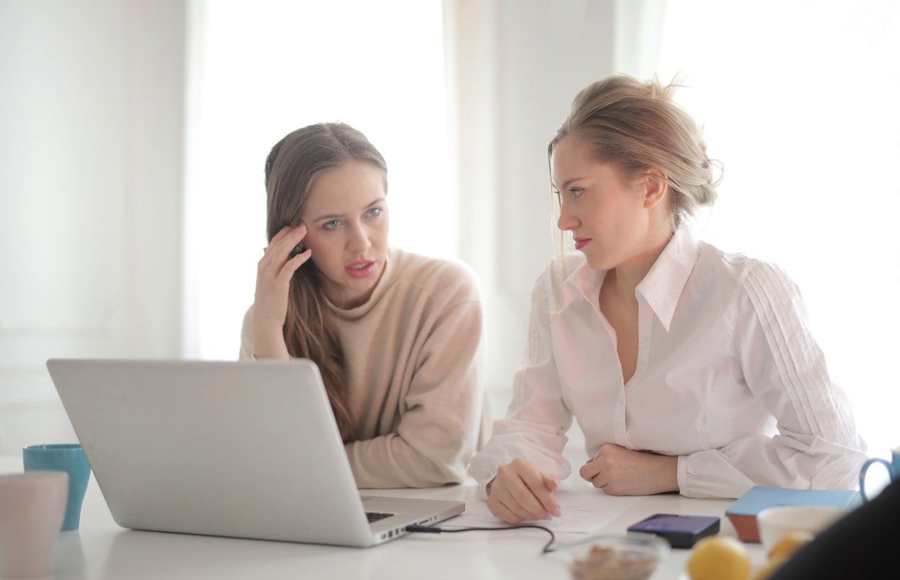 Two professionally dressed women are sitting down and talking. They are moderately stressed.