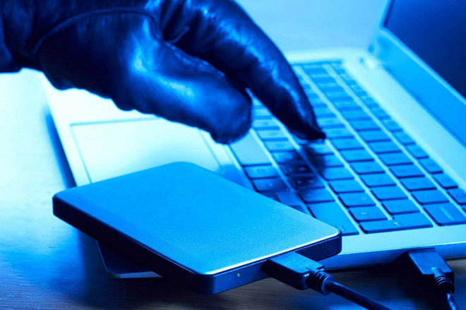 Gloved hand types on a laptop. There is an external hard drive connected to the computer, implying a malicious data theft incident